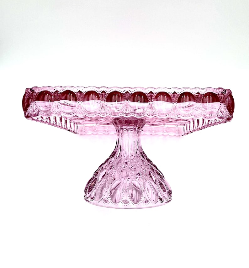 Op de afbeelding: Een roze glazen taartstandaard met een vierkante bovenkant en een decoratieve, geschulpte rand. De standaard heeft een gedetailleerde, getextureerde basis en steel. De standaard is gemaakt van glas en is ideaal voor het presenteren van taarten en andere desserts.