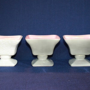 May include: Three ceramic planter pots with square bases and a pedestal stand. The pots are white and grey with a pink rim.
