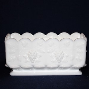 May include: White milk glass planter with a scalloped rim and a grapevine design. The planter has a square base and is approximately 15 cm wide and 10 cm tall.