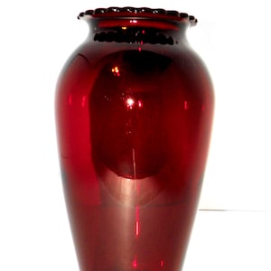 May include: A tall, ruby-red glass vase with a flared body and a ruffled rim. The vase has a glossy finish and is likely used for displaying flowers or as a decorative item. The vase is empty and stands on a white background.