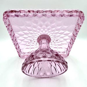 Op de afbeelding: Een roze taartstandaard in vintage-stijl met een vierkante bovenkant en een voet. De standaard heeft een gestructureerd, gewatteerd patroon op de bovenkant en decoratieve details op de voet. De randen van de bovenkant zijn geschulpt. De standaard is van glas.