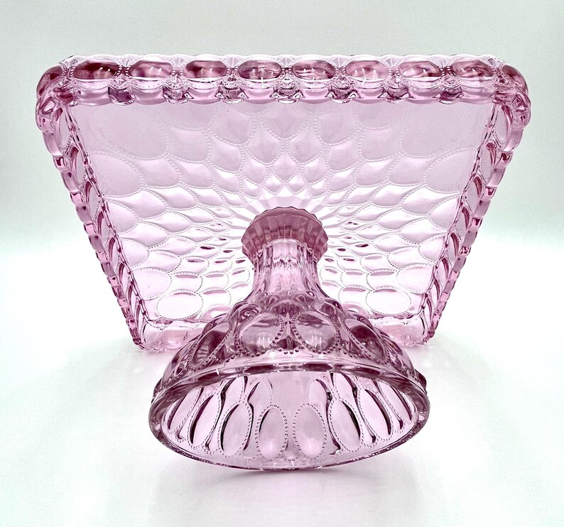 Op de afbeelding: Een roze taartstandaard in vintage-stijl met een vierkante bovenkant en een voet. De standaard heeft een gestructureerd, gewatteerd patroon op de bovenkant en decoratieve details op de voet. De randen van de bovenkant zijn geschulpt. De standaard is van glas.