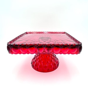 May include: A vibrant red square glass cake stand. The stand features a scalloped edge and a textured surface on the top and base. The base is a rounded shape with a textured pattern. The cake stand is a decorative piece for displaying desserts.