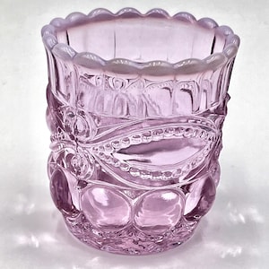 May include: A vintage, lavender-colored glass tumbler with a scalloped rim and ornate, embossed floral and geometric patterns. The glass has a rounded base and a cylindrical body, showcasing a detailed design.