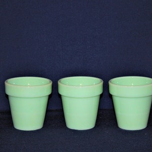 May include: Three small, light green ceramic flower pots with a slightly flared rim. The pots are cylindrical with a smooth, glossy finish. The background is a solid dark blue.