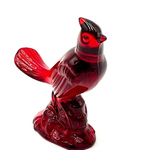 May include: A red glass cardinal figurine perched on a branch. The bird has a detailed beak and feathers.