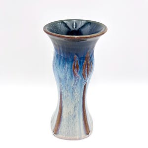 May include: A handmade ceramic vase with a flared top and a unique shape. The vase features a gradient of colors, transitioning from a dark blue rim to a light blue and white body, with brown accents. The vase is approximately 7 inches tall.