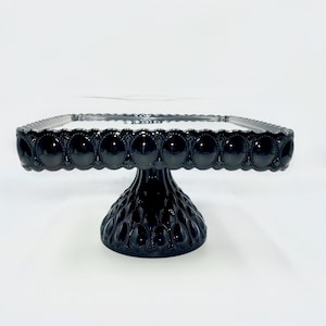May include: Black square cake stand with a mirrored top and a pedestal base. The stand has a beaded edge and a textured base.