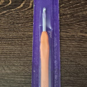 May include: An ergonomic aluminum crochet hook in a purple package. The hook has an orange handle and a silver hook end. The package text includes "5.0mm" and "1 pc/p".