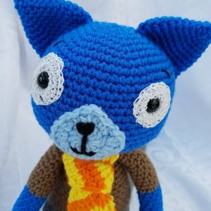 May include: Blue crocheted stuffed animal with brown and orange crocheted scarf. The animal has large white eyes with black pupils and a light blue crocheted nose.