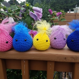 May include: Five crocheted teardrop-shaped plushies with fuzzy hair in pink, blue, yellow, purple, and blue. Each plushie has black button eyes.