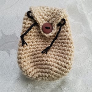 May include: A small crocheted pouch with a brown button and black drawstring closure. The pouch is made of beige yarn.