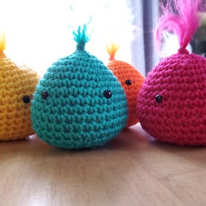 Puede incluir: Cuatro criaturas redondas de ganchillo, amarillas, turquesas, naranjas y rosas, cada una con ojos negros y un mechón de hilo. Estos objetos hechos a mano podrían ser juguetes o elementos decorativos, con un diseño caprichoso y colorido.