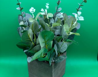 May include: A potted artificial plant with green leaves and white flowers in a gray wooden planter. The plant is on a green background.