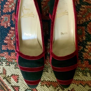 May include: A pair of black heels with red velvet trim and accents. The shoes have a pointed toe and a low heel. The interior of the shoe is beige with the text "Christian Louboutin Paris".