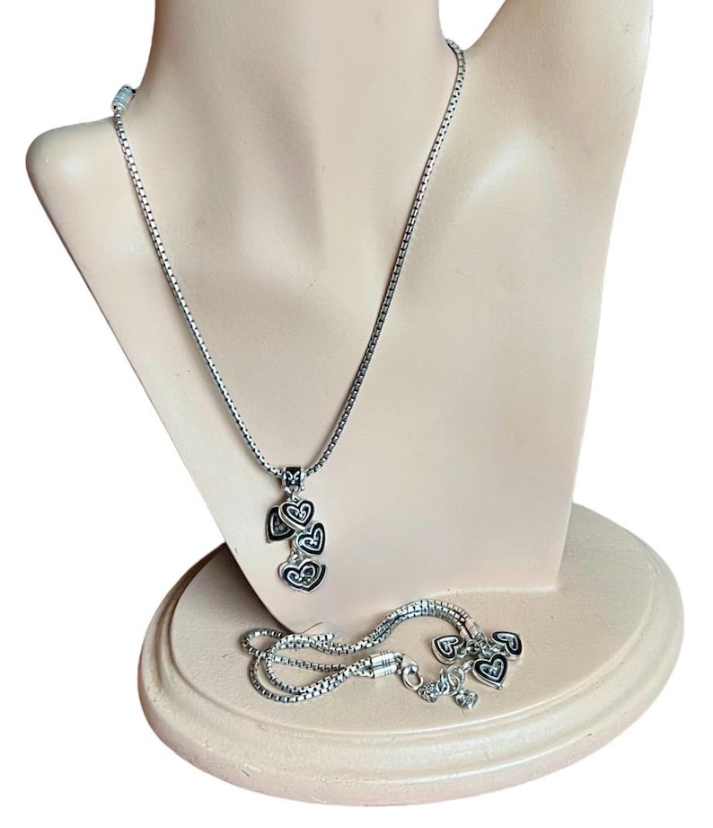 Może przedstawiać: Srebrny naszyjnik z łańcuszkiem i zawieszką w kształcie pięciu splecionych serc. Serca są czarne z srebrnymi obw&oacute;dkami. Naszyjnik znajduje się na manekinie.
