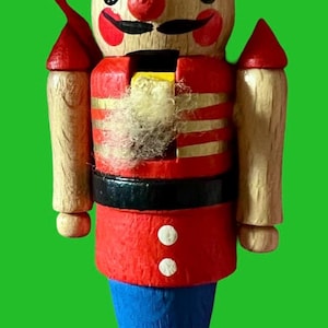 May include: A wooden nutcracker figurine with a painted design. The figurine has a red and gold jacket, blue pants, and a tall hat with blue and white stripes. A yellow ball sits atop the hat. The nutcracker has a red nose and a black mustache.