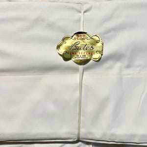 May include: A folded white sheet secured with a thin white band. A gold-colored label is attached to the center, featuring the text Bates, COMB PERCALE, and 81 x 108 BEFORE HEMMING, along with a small crown icon at the top.