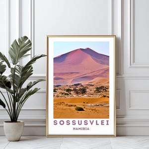 Könnte beinhalten: Gerahmter Druck von Sossusvlei, Namibia. Die Landschaft zeigt eine große rosa Sanddüne unter blauem Himmel. Der Vordergrund zeigt Wüstenvegetation und Sand. Der Druck ist in einem goldenen Rahmen, mit dem Text "Sossusvlei Namibia" am unteren Rand.