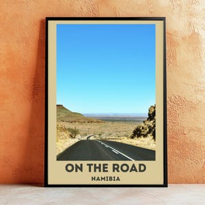 Könnte beinhalten: Gerahmtes Poster mit einer malerischen Straße, die sich durch eine Wüstenlandschaft unter einem klaren blauen Himmel schlängelt. Das Poster hat einen beigen Rand mit dem Text "ON THE ROAD NAMIBIA" am unteren Rand.