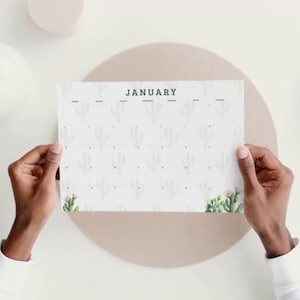 May include: A white calendar page for January, featuring a cactus pattern and held by hands. The word 'JANUARY' is printed in green. The background is a neutral beige colour.