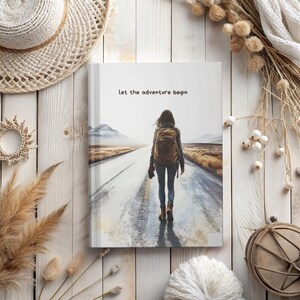 Può includere: Un diario con la scritta "Let the adventure begin" sulla copertina. L'immagine mostra una persona con uno zaino che cammina su una strada verso le montagne. Il diario è su una superficie di legno con elementi decorativi.
