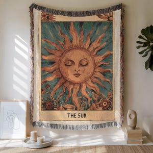 Könnte beinhalten: Gewebte Tapisserie mit einem Sonnendesign, einem ruhigen Gesicht und strahlenden Strahlen. Sie hat einen türkisfarbenen Hintergrund und einen cremefarbenen Fransenrand. Der Text "THE SUN" steht unten. Wanddekoration.