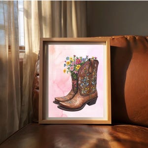 Könnte beinhalten: Ein gerahmter Druck mit einer Aquarellillustration von braunen Cowboy-Stiefeln, die mit bunten Blumen gefüllt sind. Die Stiefel haben aufwendige Blumenstickereien. Der Hintergrund ist eine weiche rosa Aquarellwäsche.