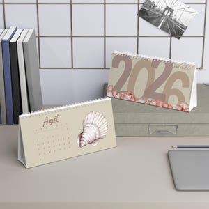 Könnte beinhalten: Zwei Tischkalender mit Spiralbindung. Ein Kalender zeigt den Monat August mit einer Muschelillustration. Der andere Kalender zeigt das Jahr 2026. Beide Kalender haben eine neutrale Farbpalette mit einem Muschelthema.