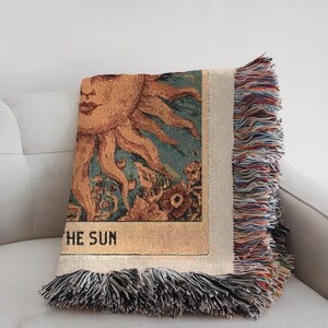 Boho woven blanket showing a tarot card in vintage style, representing "The Sun" card. Cosy throw blanket in 100% cotton, with a sunny and spiritual design in warm and natural colors. Perfect for cozy evenings at home or outdoor, available in three sizes. Must have for anyone who wants to add warm and positive decor to its home. Great gift for yourself or others.