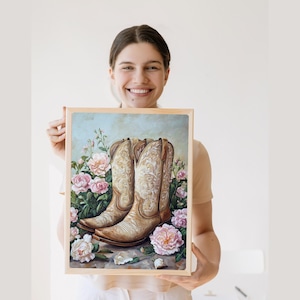 Könnte beinhalten: Gerahmtes Gemälde von hellbraunen Cowboy-Stiefeln mit aufwendigen Mustern, umgeben von rosa und weißen Rosen. Das Kunstwerk hat einen hellblauen und grünen Hintergrund und wird von einer Person gehalten.