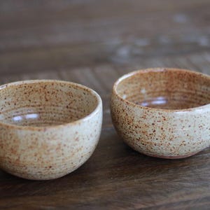 May include: Two handmade ceramic bowls. The bowls are a speckled beige color with brown speckles and a textured surface. The bowls have a rounded shape and are suitable for serving small portions of food.