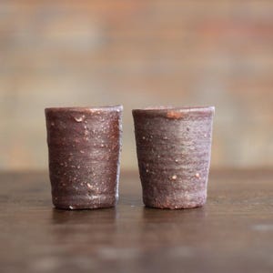 May include: Two brown ceramic cups with a speckled glaze. The cups are sitting on a wooden surface.