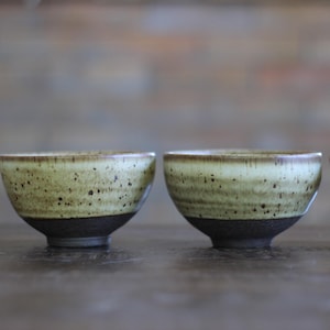 May include: A set of four ceramic bowls featuring a two-tone design with a dark, textured black base and a speckled, light olive-green upper section. The bowls have a rounded shape with a smooth rim and a distinct transition between the two colors.