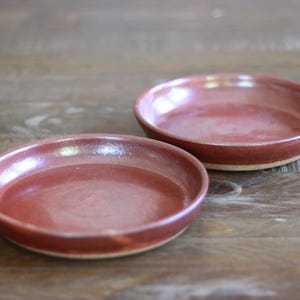 May include: Two round ceramic plates with a glossy, deep reddish-brown glaze on the interior and a smooth, curved rim. The exterior base of each plate shows a natural, unglazed tan clay texture, contrasting with the vibrant, reflective surface.