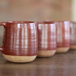 May include: A set of four ceramic mugs with a glossy, deep reddish-brown glaze on the upper portion and a textured, unglazed tan base. Each mug features a rounded body, a curved handle, and subtle vertical indentations along the exterior surface.