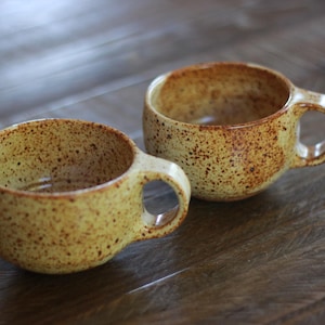 May include: Two ceramic mugs with a rounded shape and a speckled yellow and brown glaze. Each mug features a small, circular handle attached to the side. The surface of the pottery has a textured appearance with darker brown spots throughout.