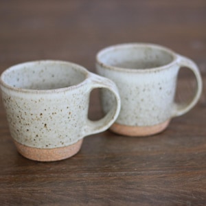 May include: Two ceramic mugs featuring a speckled off-white glaze on the upper portion and an exposed, textured terracotta-colored clay base. Each mug has a rounded handle and a slightly tapered shape, showcasing a handcrafted appearance.