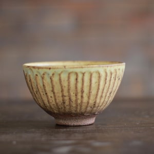 May include: A small ceramic bowl with a pale yellow glaze and vertical ribbed texture. The exterior features dark brown speckles and vertical grooves that reveal the raw, dark clay body beneath the glaze, ending in a sturdy, unglazed foot.