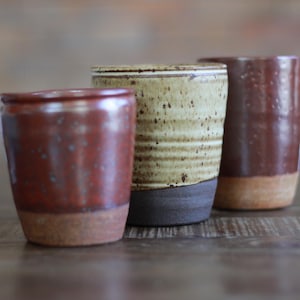 May include: Three cylindrical ceramic cups with distinct finishes. The front left cup is reddish-brown with a textured base. The center cup is light beige with horizontal ridges and a dark brown base. The back right cup is dark brown with a speckled glaze.