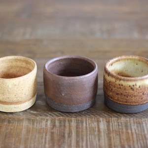 May include: Three small, cylindrical ceramic cups arranged in a row. The left cup is light beige with speckles. The middle cup is a solid dark brown with a matte finish. The right cup features a cream-colored interior and a speckled brown base.