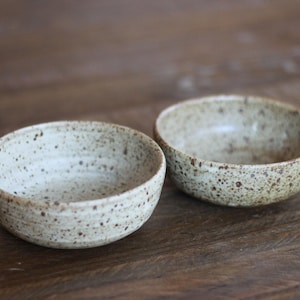 May include: Two speckled ceramic bowls. The bowls are round with a light beige color and dark brown speckles. The bowls are sitting on a wooden surface.