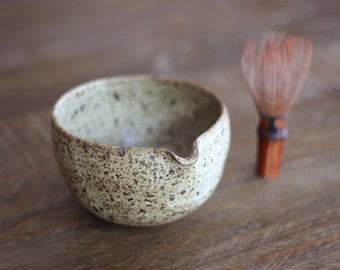 May include: A speckled light beige ceramic matcha bowl with a small pouring spout and a brown bamboo whisk. The bowl features a textured surface with dark brown flecks throughout, while the whisk has thin, light brown bristles and a handle.