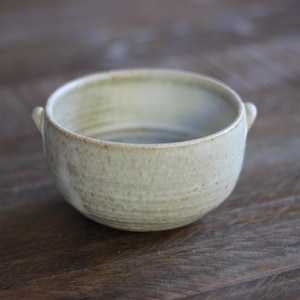 Puede incluir: Un cuenco pequeño y redondo de cerámica con un esmalte verde claro y dos pequeñas asas. El cuenco tiene una superficie texturizada y un diseño simple y rústico. Ideal para servir sopas, salsas o pequeñas porciones.