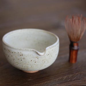 May include: A speckled, off-white ceramic tea bowl with a spout, paired with a bamboo tea whisk. The bowl has a rustic, handmade appearance, and the whisk features a dark brown handle and light brown bristles. The items are set against a blurred wooden background.