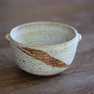 May include: A speckled ceramic bowl with a cream-colored base and a brown diagonal stripe. The bowl has two small handles and is set on a wooden surface. The bowl is likely handmade and suitable for serving food.
