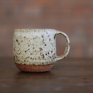 May include: A ceramic mug with a rounded body and a single handle. The upper portion features a cream-colored glaze with dark brown speckles, while the base is composed of exposed, textured reddish-brown clay, creating a distinct two-tone appearance.
