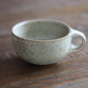 May include: A speckled, light green ceramic teacup with a rounded body and a small handle. The cup has a rustic, handmade appearance, with visible texture and subtle brown speckles. The interior is a lighter shade of green.