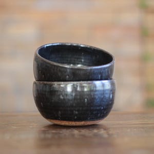May include: Two stacked ceramic bowls with a glossy, dark charcoal-black glaze featuring subtle white speckles and a visible, unglazed terracotta-colored base. The bowls have a rounded, deep shape with a smooth, reflective surface finish.