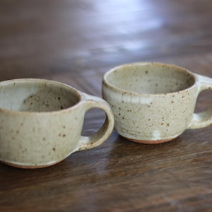 May include: Two ceramic mugs with a speckled light beige glaze and a smooth, rounded shape. Each mug features a sturdy side handle and a visible terracotta-colored base, showcasing a natural, earthy aesthetic with subtle variations in the finish.